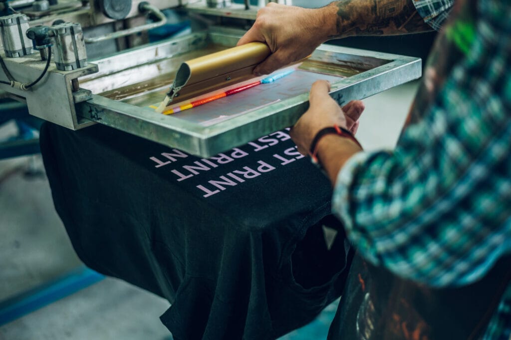 Manual screen printing process showing a test print on a black Jammy Threads t-shirt.